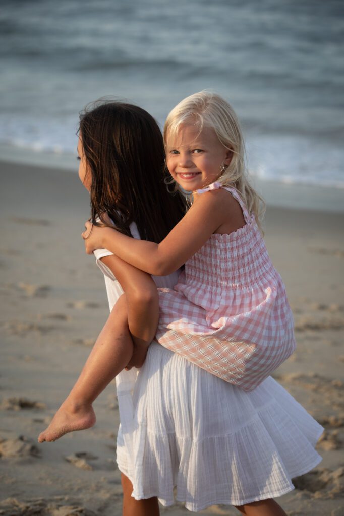 Beautiful child riding piggy back with older sibling on the beach at sunset in Duck, NC