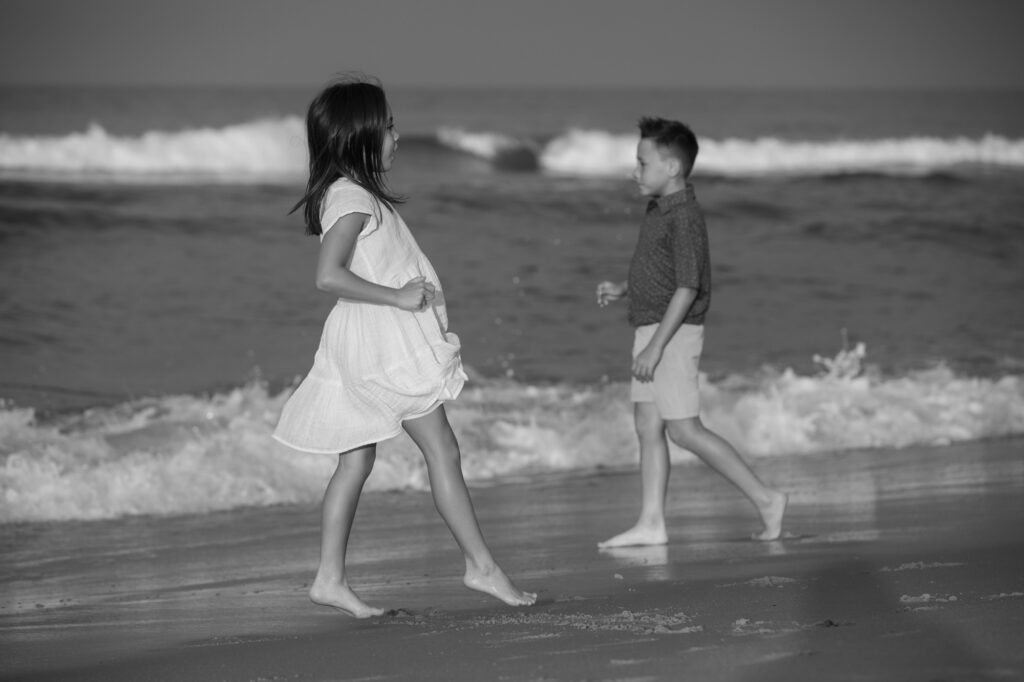 Black and white dreamy image on the beach of a girl walking opposite her brother in Duck, NC